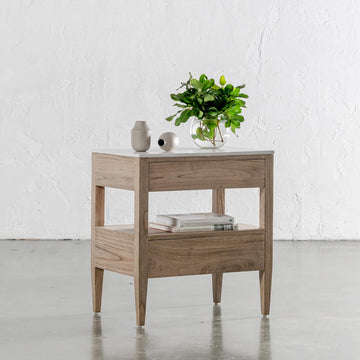 Timber bedside table featuring two drawers and a white top, styled with small ceramic vases, books, and a glass vase of green foliage.