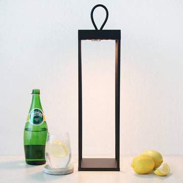 Tall, rectangular black metal LED lantern standing next to a bottle of Perrier water, a glass, and fresh lemons.