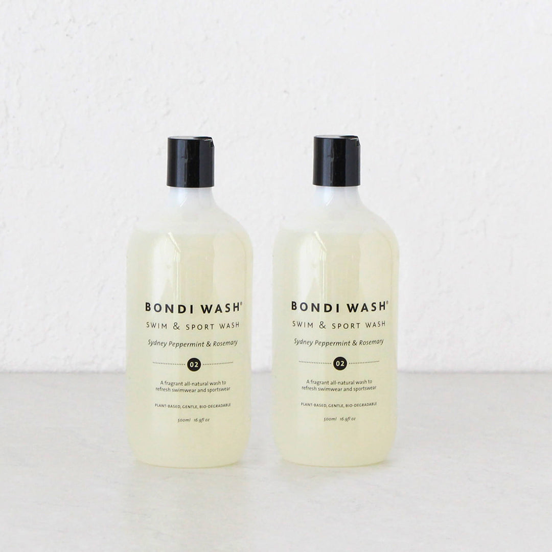 Two bottles of Bondi Wash Swim & Sport Wash in Spicy Peppermint & Rosemary scent, standing side-by-side on a white surface against a white textured wall.