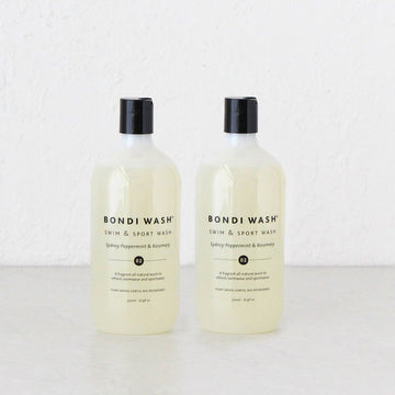 Two bottles of Bondi Wash Swim & Sport Wash in Spicy Peppermint & Rosemary scent, standing side-by-side on a white surface against a white textured wall.