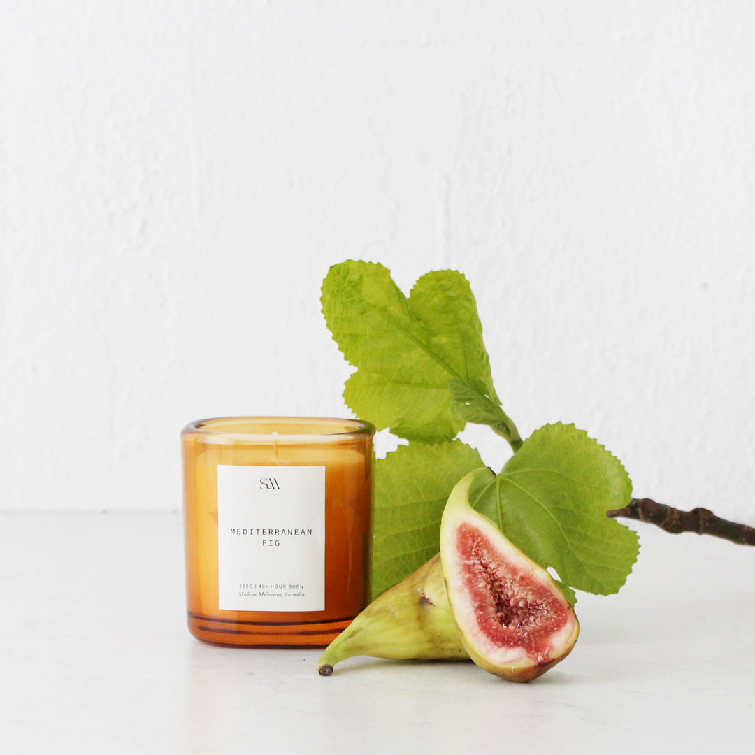 A lit Mediterranean Fig scented candle in an amber glass jar, styled with a fresh halved fig and green leaves.