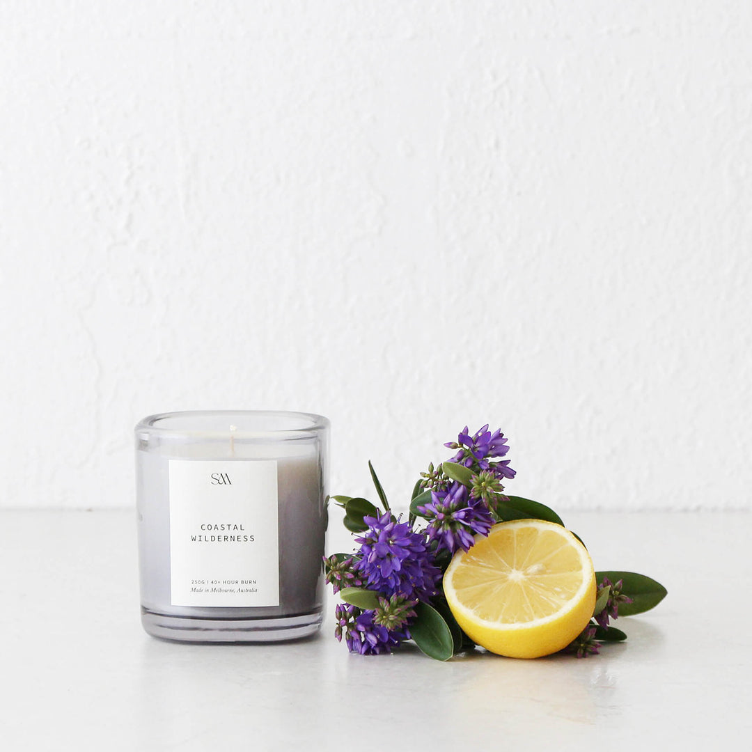 A white speckled ceramic footed display stand holding a rustic bowl, a large candle, essential oil, and decorative branches against a white wall.