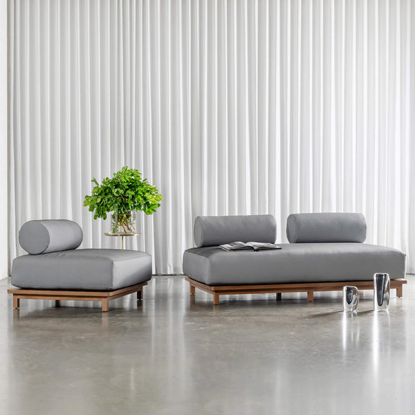 A modern grey outdoor lounge sofa and ottoman with cylindrical backrests and wooden bases, styled indoors on a polished concrete floor.