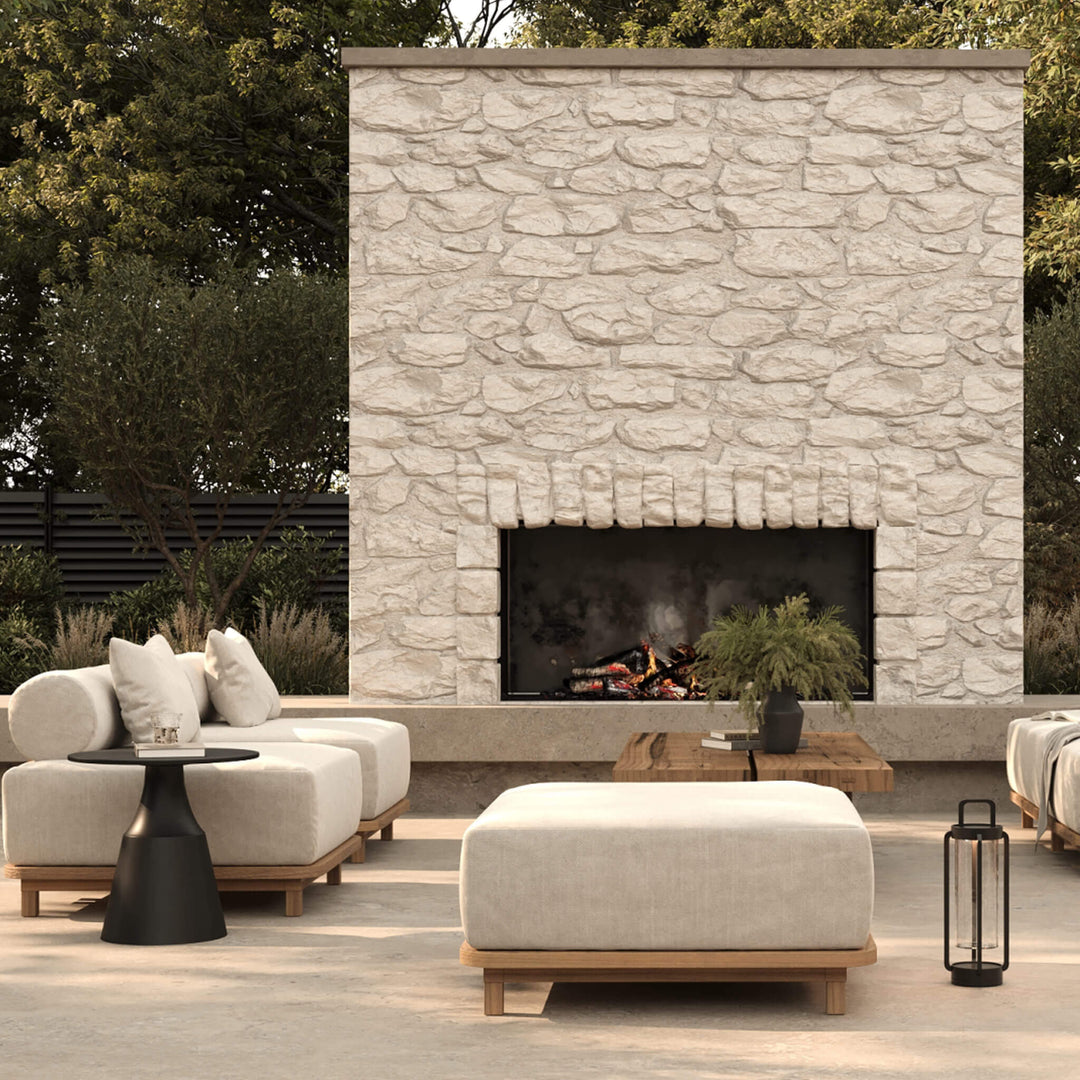 Outdoor living area featuring a large stone fireplace, two modular cream lounge chairs, an ottoman, and a timber coffee table.